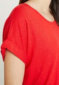 Top rosso a maniche corte con scollatura a barca, caratterizzato da risvolti e un tessuto morbido e testurizzato. Dettagli delle cuciture lungo la spalla.