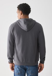 Man with short curly hair wearing a dark gray hoodie and blue jeans, shown from the back against a plain white background.