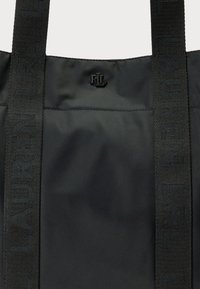 Black nylon tote bag featuring contrasting black logo hardware and textured straps with raised lettering. Smooth surface, minimalistic design.