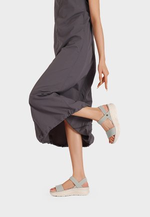 Donna che indossa pantaloni grigi a gamba larga, solleva una gamba mostrando sandali grigio chiaro con suole spesse e cinturini regolabili.