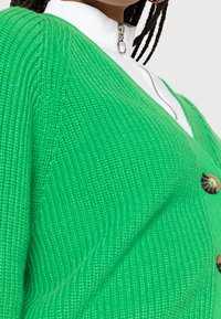 Grön stickad cardigan med V-ringning, ribbad textur och bruna knappar. Lager på en vit zip-up topp.