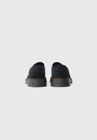 Chaussures slip-on en suède bleu foncé avec une semelle en caoutchouc noir bas et des détails de couture subtils à l'arrière. Design minimaliste sans marque visible.