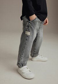 Jean en denim gris avec une coupe ample, dotés de poches latérales et d'une étiquette sur la poche arrière, associés à des baskets blanches à Velcro.
