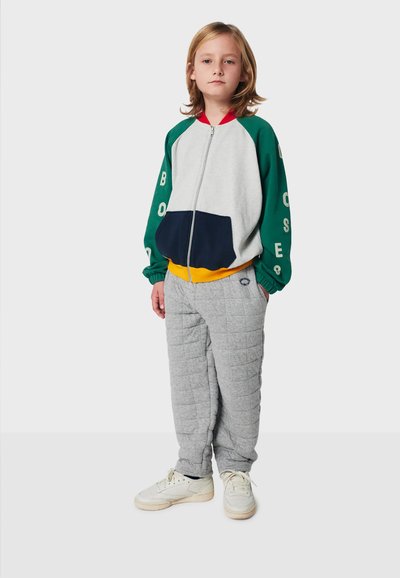 Färgglad zip-up jacka med grå kropp, gröna ärmar och marinblå ficka. Grå vadderade byxor med strukturerad mönster. Vita sneakers fulländar looken.