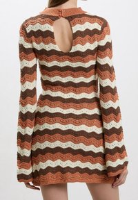 Femme portant une robe en maille à manches longues avec des rayures horizontales ondulées en rouille, marron et crème, dotée d'une ouverture en forme de goutte d'eau dans le dos avec une fermeture par bouton.