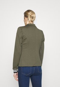 Veste ajustée vert olive avec col, conçue avec des coutures courbées. Poignets noirs avec des rayures blanches et texture de tissu lisse.