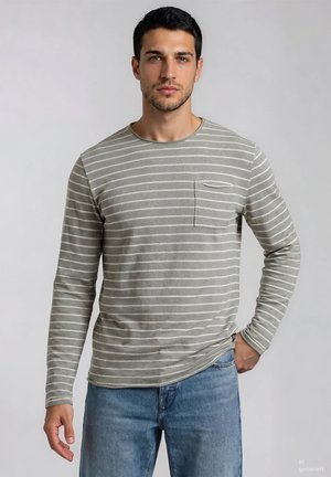 Hombre con camisa gris de manga larga con rayas blancas y bolsillo en el pecho, combinado con vaqueros azules, de pie frente a un fondo liso y claro.