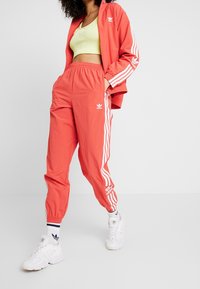 Červená tepláková souprava s bílým designem tří pruhů, lehká tkanina, elastický pas a odpovídající krátký top. Bílé tenisky a ponožky.