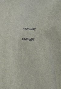 Grön tyg med två svarta tryckta "SAMSØE"-logotyper, en ovanför den andra, centrerade och något förskjutna.