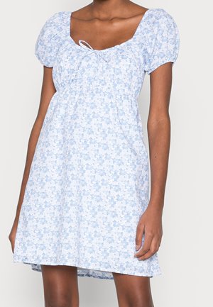 Femme portant une robe courte fleurie bleu clair avec des manches bouffantes et un col noué, debout devant un fond uni.