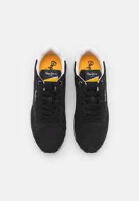 Chaussures de sport noires avec une tige en tissu texturé, accents blancs et lacets noirs. Semelle intérieure jaune avec détail du logo. Design léger.