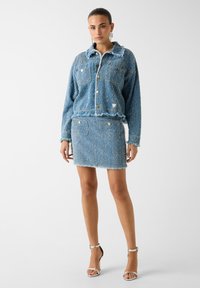 Giacca di jeans blu con bordi sfrangiati e tasca sul petto, abbinata a una gonna coordinata con bordi sfrangiati. Inserti con bottoni argentati completano il look.