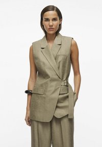 Blazer sin mangas beige con diseño envuelto, un bolsillo y detalle de cinturón. Acompañado de pantalones sueltos beige, de tela texturizada.