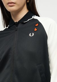 Femme portant une veste zippée noire et blanche avec un logo de laurier blanc et deux petites patchs en forme de cœur rouge sur la poitrine.