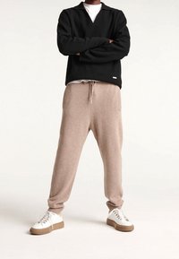 Schwarzer Pullover mit langen Ärmeln und V-Ausschnitt; beiger Strick-Jogginghose mit Kordelzug und Seitentaschen; weiße Sneakers mit sandfarbenen Akzenten.