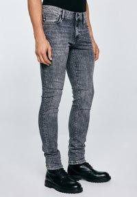 Isik, kes kannab kehakinnitusega halli happepesuga teksaseid ja musti pits-up nahksaapaid, seisab ühevärvilise heleda tausta ees.