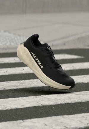 Zapatilla de correr negra con suela blanca y marca "ALTRA" en el aire sobre un paso de peatones con gotas de agua salpicando.