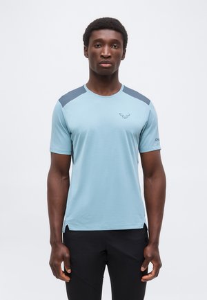 Bărbat stând cu fața înainte, purtând un tricou sport bleu deschis cu panouri închise la umăr și pantaloni negri, pe un fundal simplu.