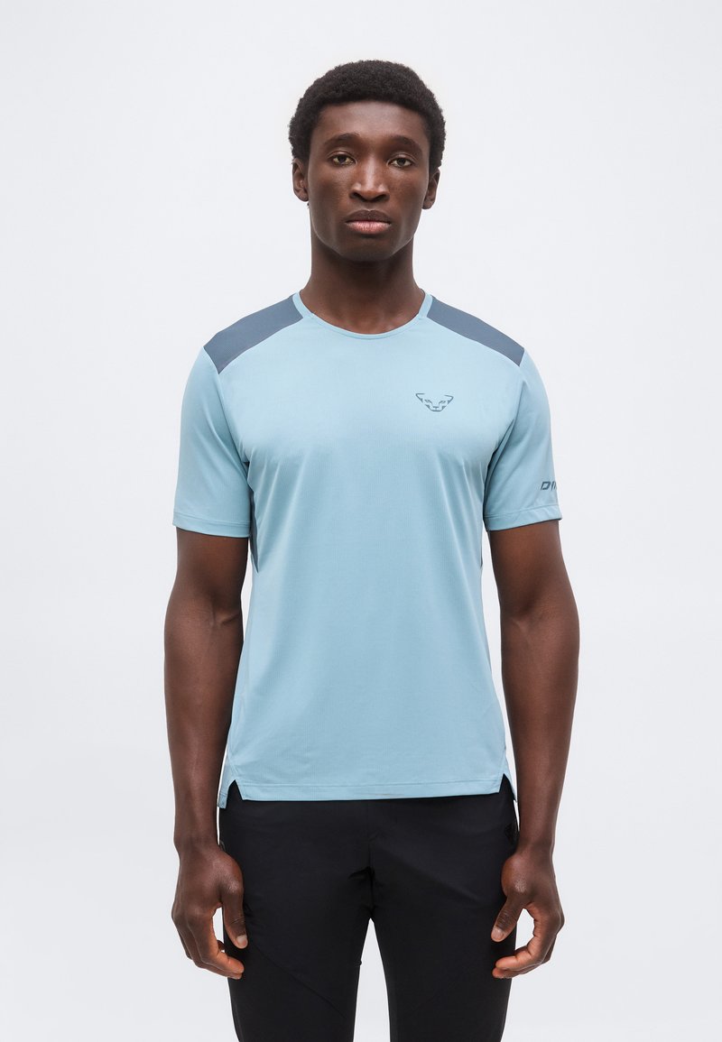 Άνδρας που στέκεται με το πρόσωπο προς τα εμπρός, φοράει ένα ανοιχτό μπλε αθλητικό πουκάμισο με σκούρα πάνελ στους ώμους και μαύρο παντελόνι ενάντια σε μια απλή φόντο.