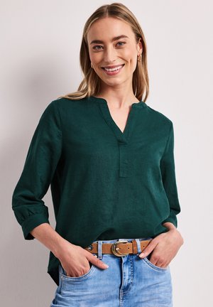 Kobieta uśmiechnięta, ubrana w ciemnozieloną bluzkę z długim rękawem włożoną do jasnoniebieskich dżinsów z brązowym paskiem, ręce w kieszeniach, jasne tło.