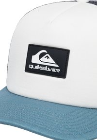 Hvid baseballkasket med en blå skygge, der har et sort rektangulært patch med Quiksilver-logoet og bjerggrafik. Tekstureret stof.