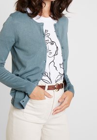 Lichtblauwe vest van zacht materiaal, met een ronde hals, knoopsluiting aan de voorkant en geribbelde manchetten. Draagt over een wit T-shirt met opdruk en beige broek.