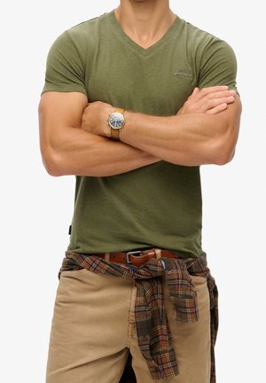 Olivgrünes T-Shirt mit V-Ausschnitt, kurze Ärmel, getragen mit beigen Hosen und einem karierten Hemd, das in der Taille gebunden ist. Braune Uhr am Handgelenk, muskulöse Arme verschränkt.