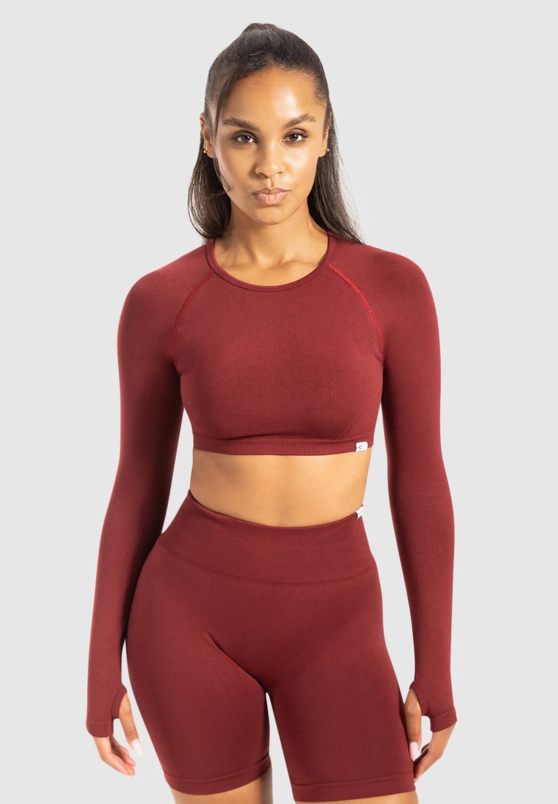 Vrouw staat met een bijpassende, getailleerde bordeauxrode langharige crop top en hoge fietsenbroek tegen een effen achtergrond.