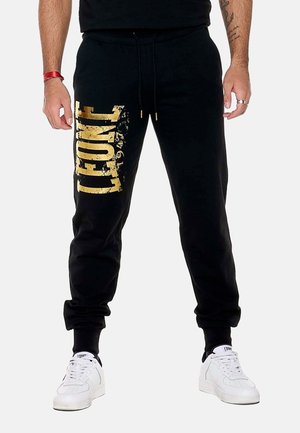 Homme portant un pantalon de jogging noir avec un grand texte doré "FONE" sur la jambe gauche et des baskets blanches sur un fond uni.