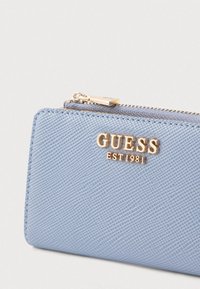 Világoskék cipzáras pénztárca texturált felülettel, aranyszínű "GUESS EST1981" logóval az elején.