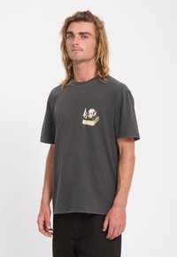 Jeune homme aux longs cheveux portant un t-shirt gris foncé avec un petit dessin de crâne et de livre sur la poitrine gauche, debout devant un fond uni.