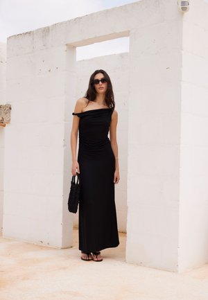 Kvinde iført lang sort off-shoulder kjole, sorte solbriller og sandaler, stående i hvidt minimalistisk arkitektonisk miljø med en sort flettet håndtaske.