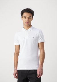 Valkoinen polo-paita, joka on valmistettu puuvillasta, kauluksen omaava, lyhythihainen ja jossa on pieni musta logo vasemmassa rinnassa.