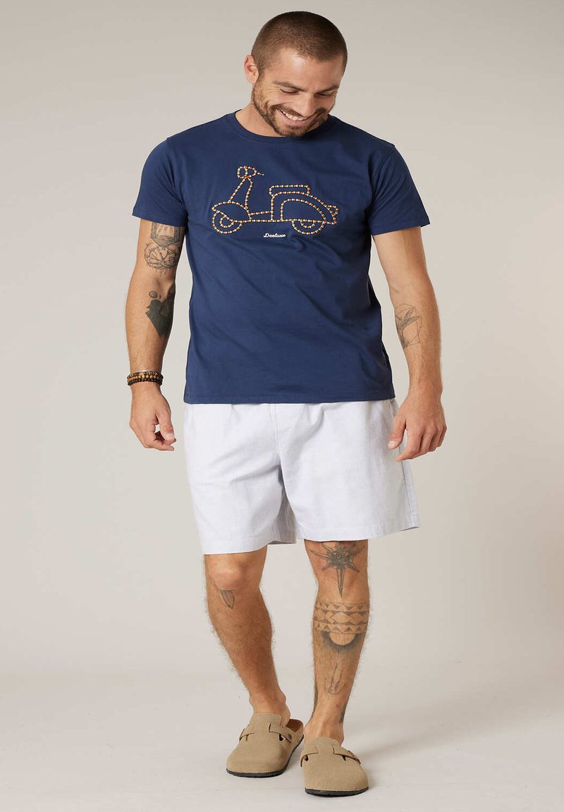 Homme souriant avec des tatouages, portant un t-shirt bleu marine avec un dessin de scooter orange, un short blanc, des chaussures marron sans lacets et des bracelets en perles.
