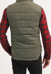 Olivgrüwest gepolsterte Weste mit hohem Kragen, ausgestattet mit horizontalen Nähten und einer strukturierten Oberfläche, getragen über einem rot-schwarz karierten Hemd.