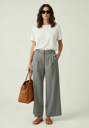 Femme portant des lunettes de soleil noires, chemise blanche à manches courtes, pantalon gris à jambes larges, sandales marron, tenant un grand sac à main marron.