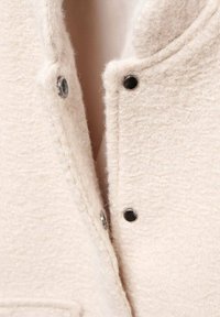 Light beige wool jacket with a textured surface, featuring black snap fasteners and a stand collar. Close-up view of the front opening.