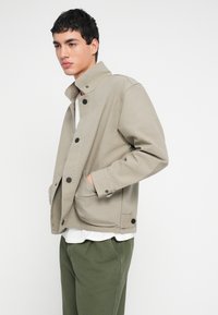 Bež platnena jakna z ovratnikom v stoječem ovratniku, črnimi gumbi in sprednjimi žepi. Nošena prek bele srajce in zelenih hlač, z razigranim prileganjem.