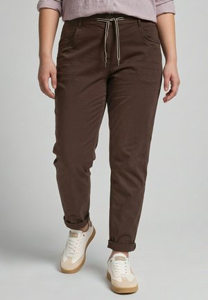 Personne portant un pantalon marron foncé retroussé avec une taille à cordon et des baskets blanches, debout devant un fond uni.