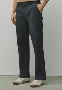 Homme portant un pantalon noir droit et des baskets blanches, debout avec une main dans la poche devant un mur uni.
