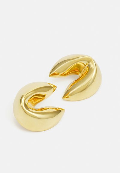 Náušnice s klipem z kovu ve zlatém odstínu s hladkým, reflexním povrchem; mají zakřivený tvar připomínající drápy s rozděleným designem.