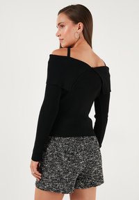 Žena s dugom smešnom kosom nosi crni pulover bez ramena s rebrastim uzorkom i teksturirane crno-bijele kratke hlače, gleda u stranu naspram jednostavne pozadine.