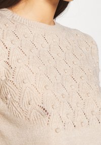 Beige gebreide trui met een textuurpatroon van verhoogde stippen, ronde halslijn en ribbelboord. De stof ziet er zacht en lichtgewicht uit.