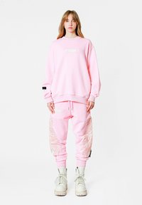 Sudadera rosa con cuello redondo y etiqueta frontal, combinada con joggers a juego que presentan diseños de alas bordadas y puños elásticos.
