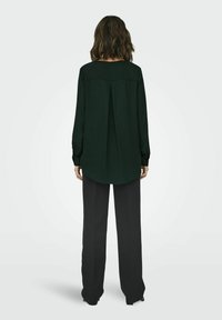 Blusa verde scuro a maniche lunghe con scollatura rotonda e piega sul retro, abbinata a pantaloni neri a vita alta e gamba larga.