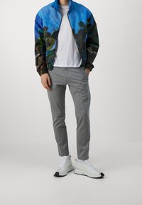 Bomberjacka med ett blått och grönt abstrakt mönster, vit t-shirt, grå slim-fit byxor och vita sneakers med svarta accenter.