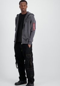 Felpa grigia con zip e tasche frontali, abbinata a pantaloni cargo neri con più tasche e cucitura gialla. Sneakers nere.