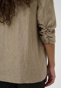 Blazer texturé beige clair, avec une finition froissée et des manches froncées. Le tissu a un léger éclat et est taillé pour un ajustement ample.