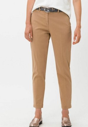 Kvinde iført beige, tætsiddende ankel-lange bukser, hvid bluse, sort mønstret bælte og metalliske loafers stående mod en hvid baggrund.