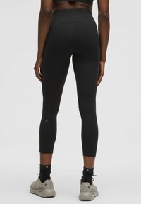 Leggings neri a vita alta con design senza cuciture, lunghezza cropped e tasche posteriori. Indossati con scarpe da ginnastica di colore neutro e calze nere.